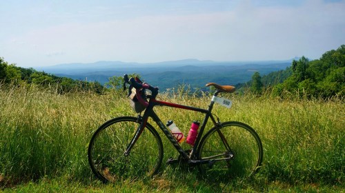 A bicycle rests in a grassy field, overlooking a scenic mountain landscape under a clear blue sky.