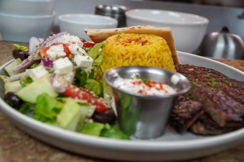 A plate featuring grilled steak, yellow rice, a fresh salad, and a small cup of sauce, set in a restaurant kitchen.