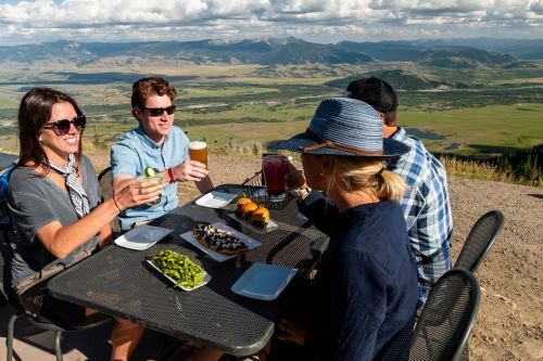 A group of four friends enjoying drinks and sushi at an outdoor table with a scenic mountain view.