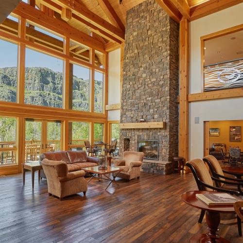 Spacious lodge interior with wooden beams, stone fireplace, and large windows showcasing a scenic view. Cozy seating area.