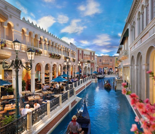 A picturesque canal scene with gondolas, restaurants, and ornate architecture under a blue sky.