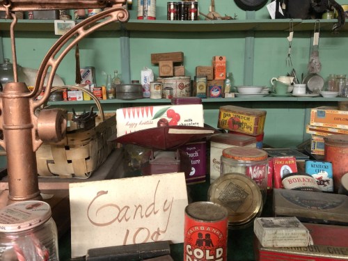 A vintage display of assorted old-fashioned goods, including candy, jars, and kitchenware on a green shelf.