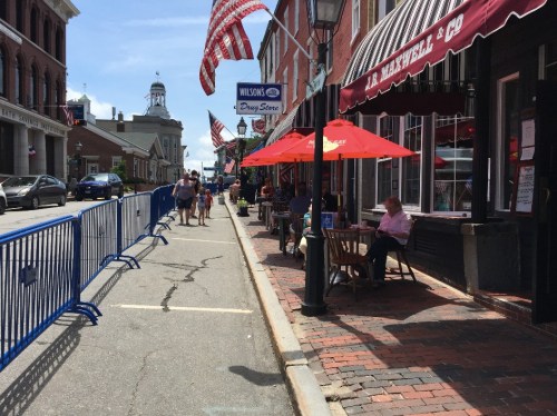 A street scene with outdoor dining, umbrellas, and American flags, featuring people walking and barriers along the sidewalk.
