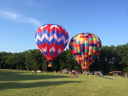 Two colorful hot air balloons are inflated and ready for flight in a grassy field under a clear blue sky.