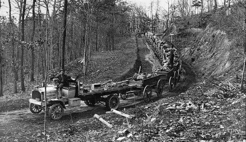 A vintage truck transporting logs along a dirt road, with workers and equipment in a wooded area.