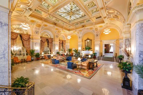 Elegant hotel lobby with ornate ceilings, plush seating, and decorative plants, featuring a grand fireplace and large windows.