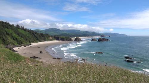 A scenic coastal view with sandy beach, waves, and lush green hills under a blue sky with scattered clouds.