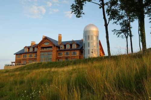 A large wooden lodge with a modern silvery silo, surrounded by tall grass and trees under a clear sky.