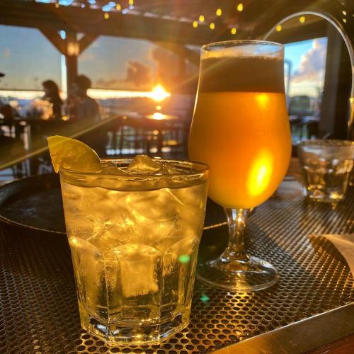 A glass of iced drink with a lime slice and a tall beer glass, with a sunset view in the background.