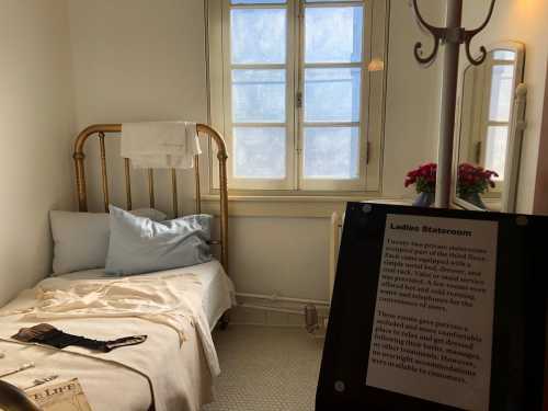 A small, vintage bedroom with a bed, light blue bedding, and a sign labeled "Ladies Stateroom" beside a window.