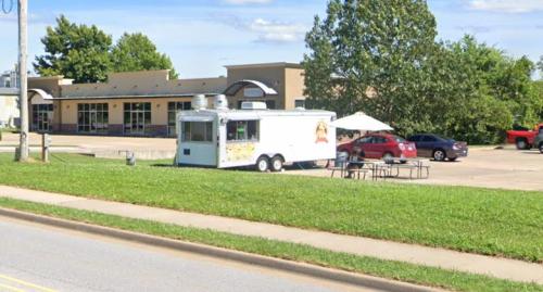 A food truck parked on grass near a building, with tables and chairs set up outside under a large umbrella.