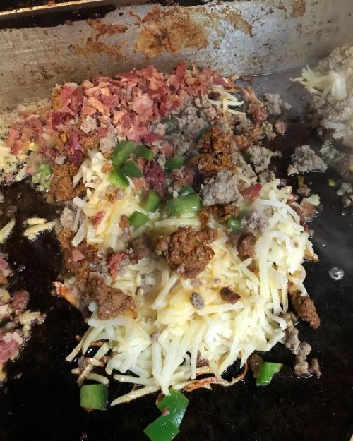 Sizzling hash browns topped with ground beef, bacon bits, and green peppers on a griddle. Steam rising from the food.