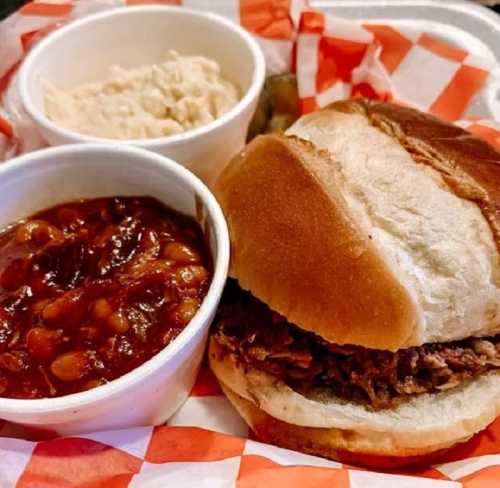 A plate featuring a sandwich with pulled meat, a side of baked beans, and a cup of coleslaw on a checkered paper background.