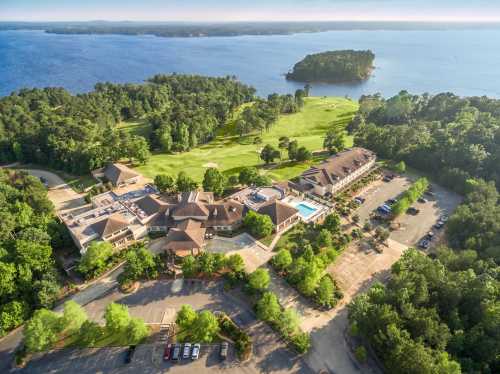 Aerial view of a resort near a lake, featuring a golf course, buildings, and parking areas surrounded by trees.