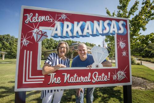 Two smiling people pose in front of a colorful sign that reads "Helena, Arkansas - The Natural State."