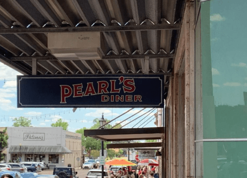 Sign for Pearl's Diner hanging above a street with outdoor seating and shops in the background.