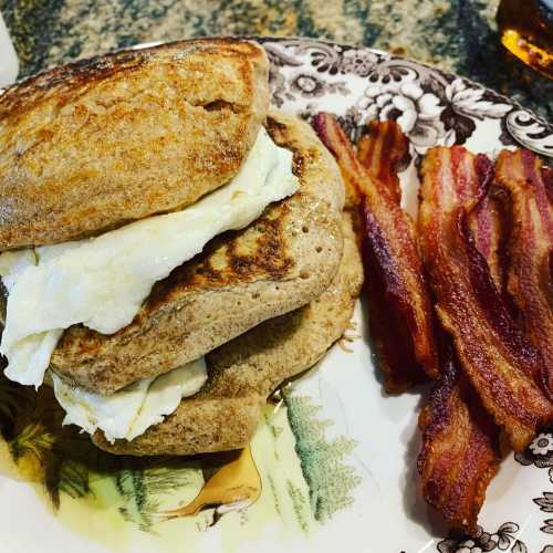 A plate with a stacked sandwich of eggs on toasted bread, accompanied by crispy bacon strips.