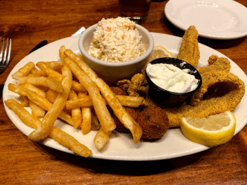 A plate of fried fish, coleslaw, fries, and dipping sauce, garnished with lemon wedges.
