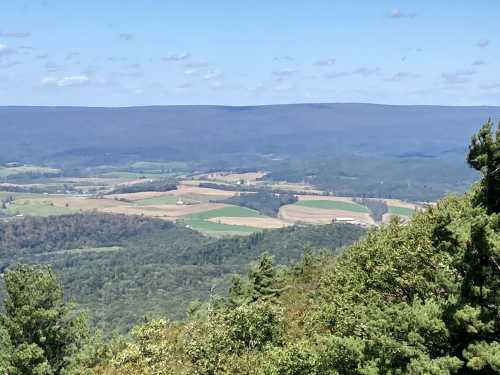 A panoramic view of rolling hills and farmland under a clear blue sky, with scattered clouds in the distance.