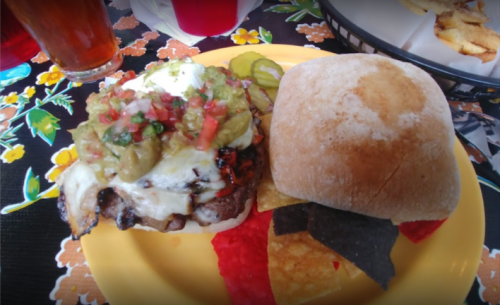 A hearty burger topped with salsa, jalapeños, and cheese, served with tortilla chips on a colorful tablecloth.