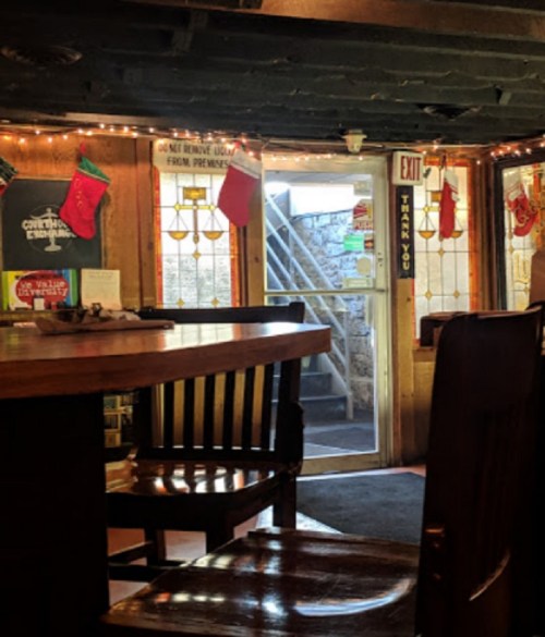 A cozy interior of a rustic bar with wooden furniture, festive decorations, and a staircase in the background.