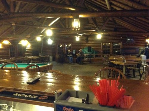 Interior of a rustic log cabin bar with wooden beams, a pool table, and two people seated in the background.