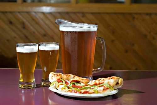 A pitcher of beer with two glasses and a plate of pizza topped with green peppers on a table.