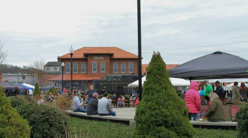 A bustling outdoor market with people, tents, and a historic train station in the background under a cloudy sky.