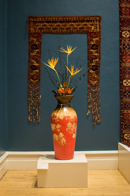 A decorative vase with bird of paradise flowers, displayed against a blue wall and ornate textile hangings.