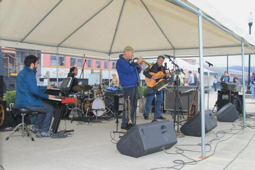 A band performs under a tent, featuring a trombone player, guitarist, drummer, and keyboardist at an outdoor event.