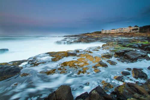 A rocky shoreline with gentle waves, a cloudy sky, and a distant building along the coast.