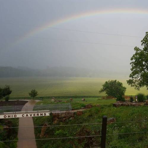 A misty landscape with a rainbow arching over green fields and trees, near the Ohio Caverns entrance.