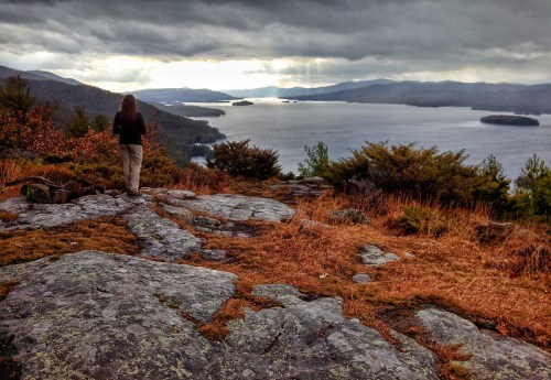 A person stands on rocky terrain, overlooking a lake surrounded by mountains under a cloudy sky.