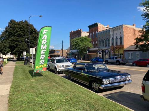 A classic car parked on a street beside a green "Farmers Market" banner and historic buildings in the background.