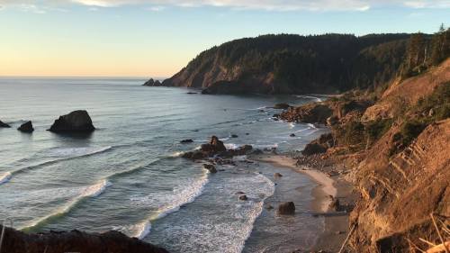 A serene coastal view at sunset, featuring waves, rocky shores, and lush green hills in the background.