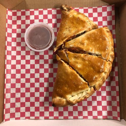 A golden-brown calzone cut into slices, served with a small cup of marinara sauce, on a red and white checkered box.