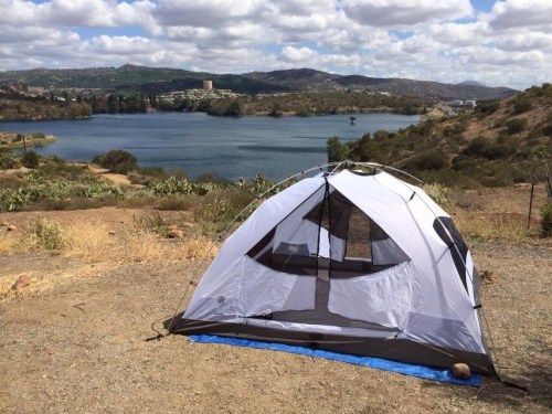 A tent set up on dry ground near a lake, with hills and clouds in the background.