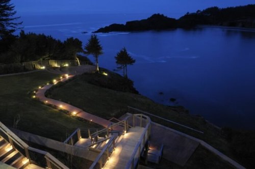 A serene coastal view at dusk, featuring a lit pathway winding through lush greenery towards the ocean.