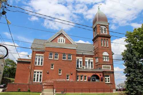 Historic red brick building with a tower, surrounded by trees and power lines, under a blue sky with clouds.