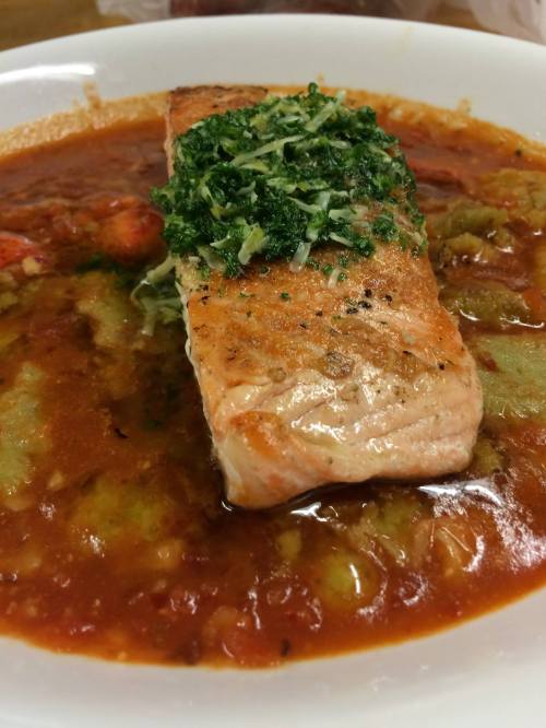 A piece of grilled salmon topped with green herbs, served in a rich tomato-based sauce with vegetables.