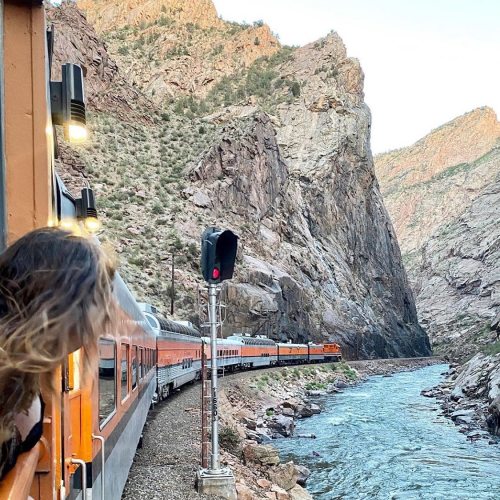 A train travels alongside a river through a rocky canyon, with a person looking out from the train window.