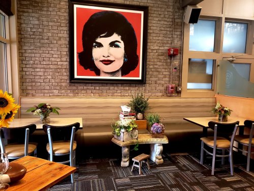 A cozy restaurant interior featuring wooden tables, a large portrait on the wall, and decorative plants.