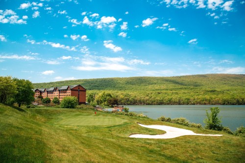 A scenic view of a lakeside resort with green hills, blue sky, and a golf course in the foreground.