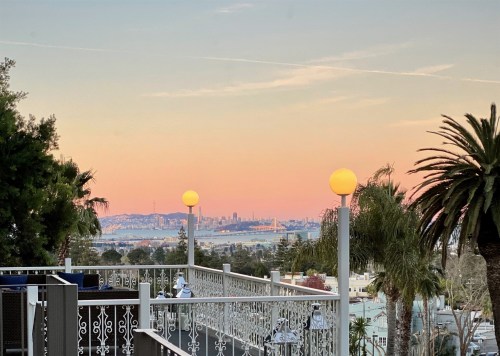 A scenic view of a city skyline at sunset, framed by palm trees and glowing lamps on a balcony.
