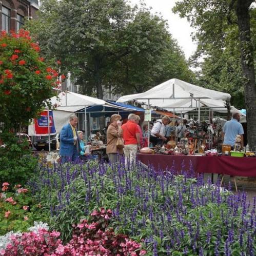 A bustling outdoor market with vendors under tents, surrounded by colorful flowers and trees.