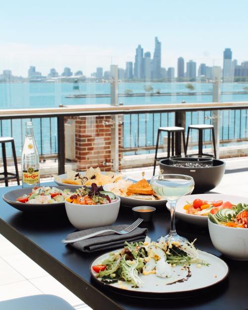 A table with various colorful dishes and drinks, overlooking a city skyline by the water on a sunny day.