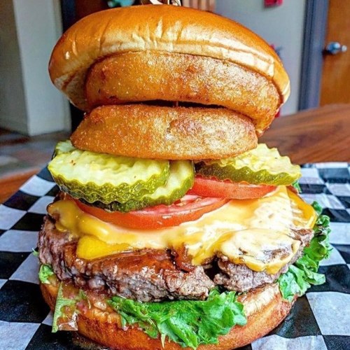 A towering burger with a beef patty, melted cheese, lettuce, tomato, pickles, and onion rings on a toasted bun.
