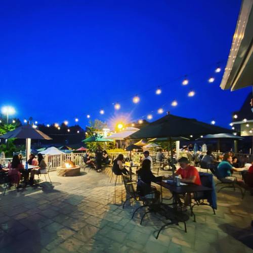 Outdoor dining area at night, with string lights and umbrellas, featuring people enjoying meals and a cozy atmosphere.