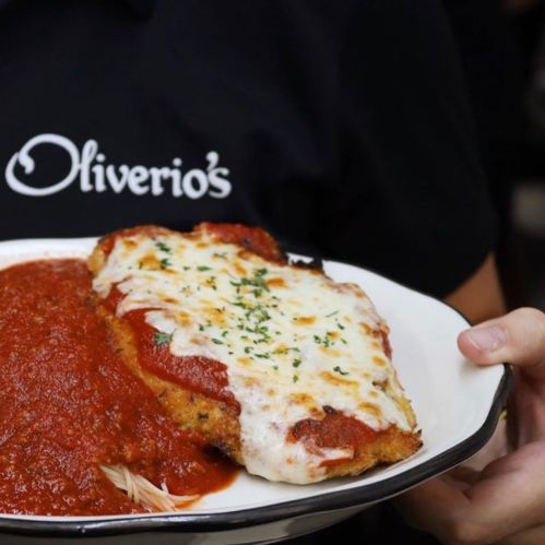 A person holds a plate of chicken parmesan with marinara sauce and pasta, wearing an "Oliverio's" shirt.