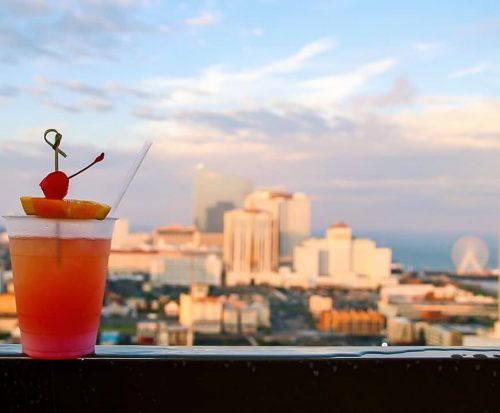 A colorful cocktail with a cherry and lemon slice, set against a blurred city skyline and sunset.